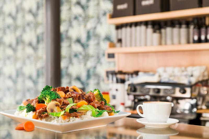 Prato de carne com legumes e arroz servido ao lado de uma chávena de café, em cima de uma montra de vidro